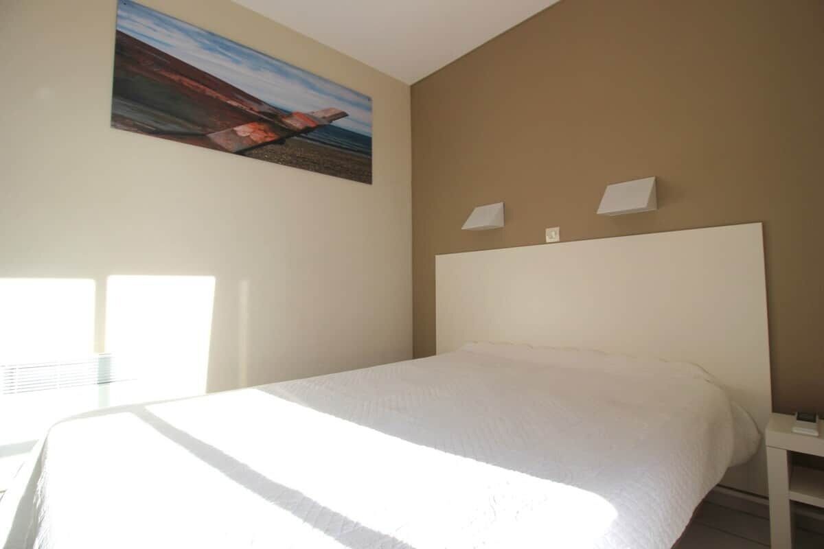A hotel room with a large bed, a painting on the wall, and a window providing natural light.