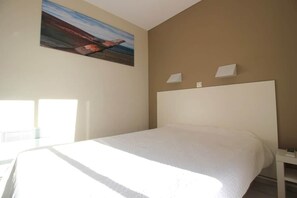 A hotel room with a large bed, a painting on the wall, and a window providing natural light.