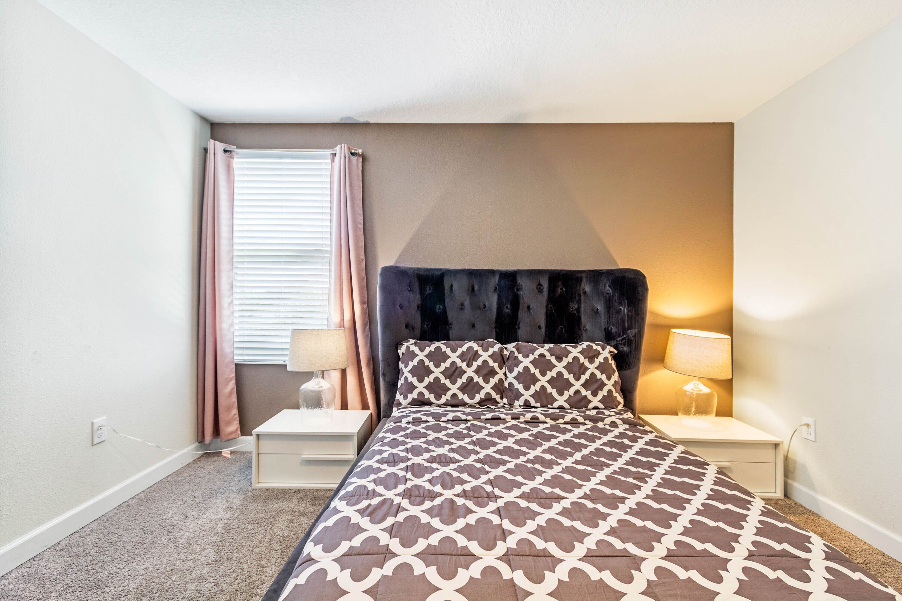 Une chambre à coucher comprenant un lit, deux tables de chevet, une commode, une lampe, une fenêtre avec des rideaux et un luminaire fixé au mur.