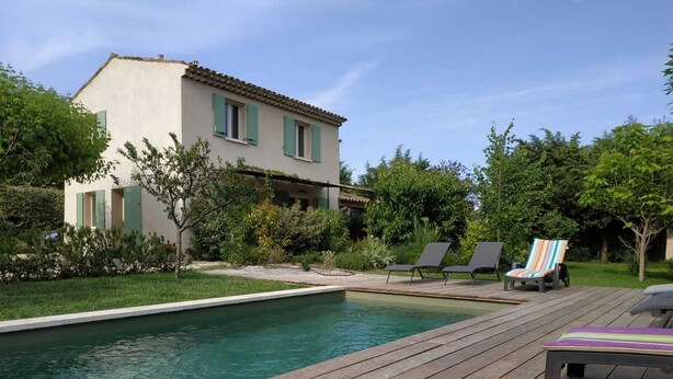 A pool area with a wooden deck, lounge chairs, and a house in the background.