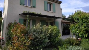 A two-story house with green shutters, a covered porch, and a garden with various plants and shrubs.