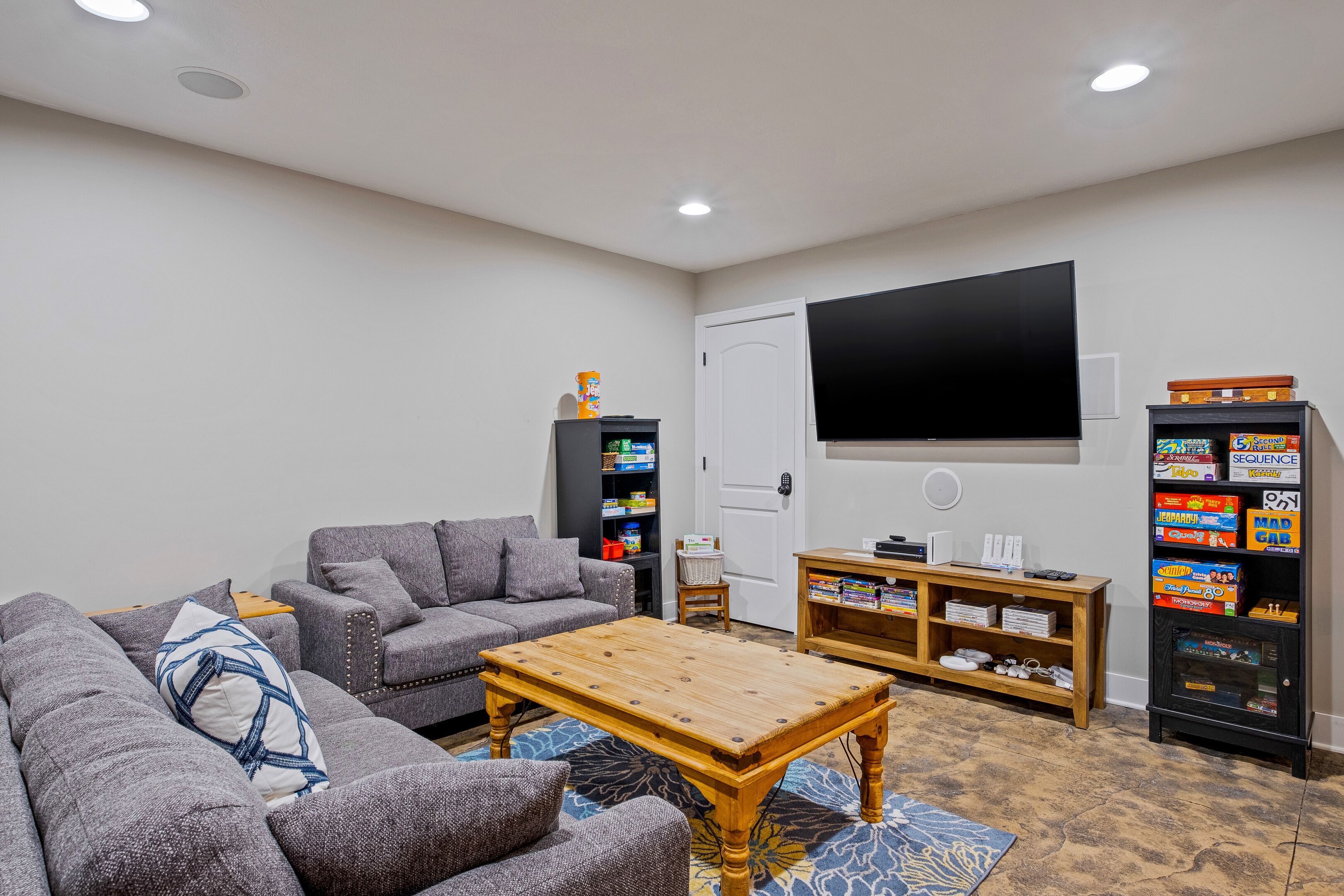 A living room with a gray sofa, a wooden coffee table, a TV mounted on the wall, and a bookshelf with video games.