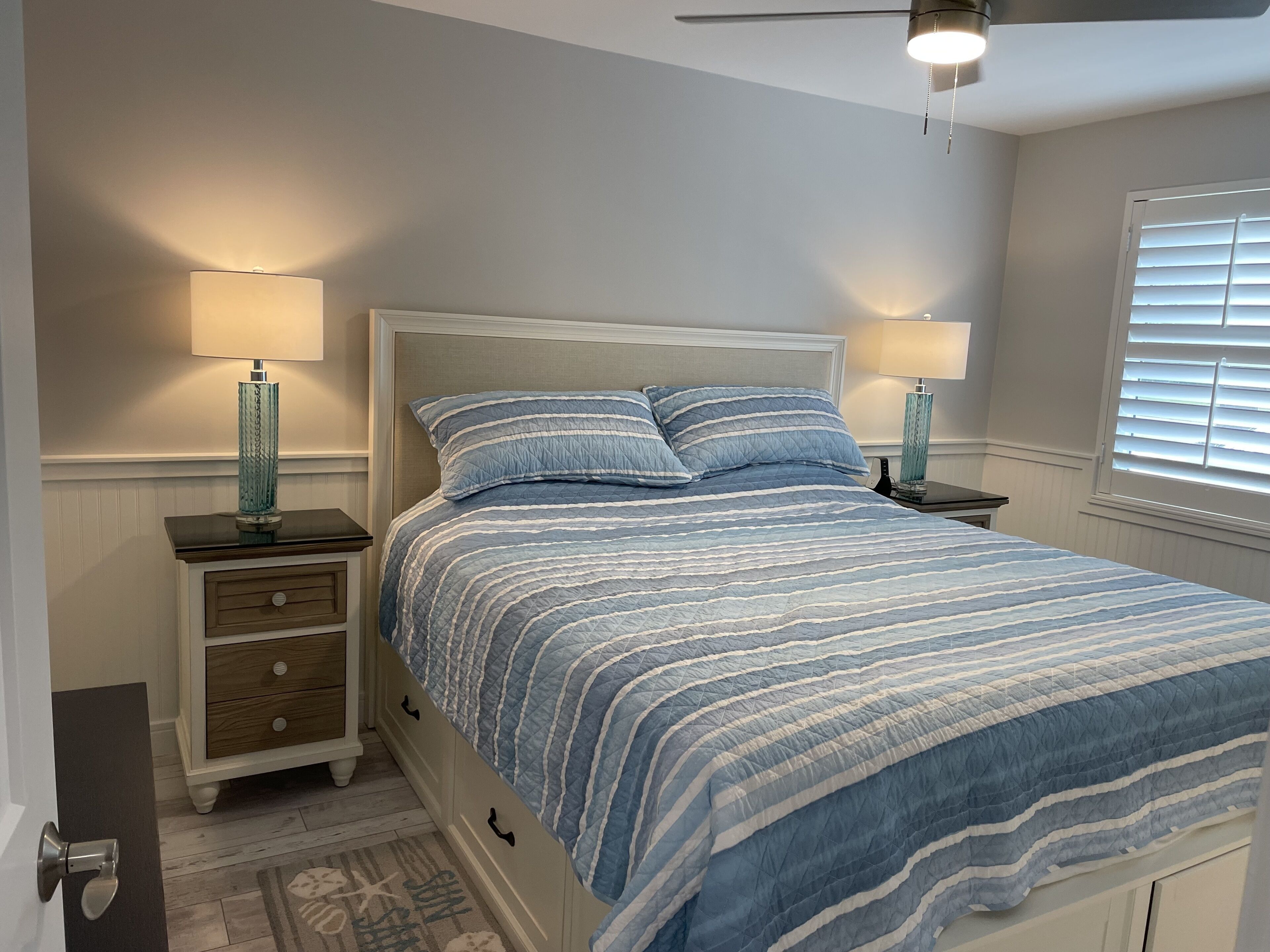 A bedroom with a bed, two bedside tables, a nightstand, a door, and a window with blinds.