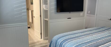 Ein Schlafzimmer mit Bett, einem an der Wand befestigten Fernseher, einer Tür zu einem Badezimmer und einem Flur.