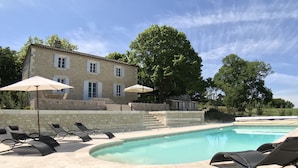 Ein Poolbereich mit Liegestühlen und Sonnenschirmen, ein Steinhaus und ein großer Baum.
