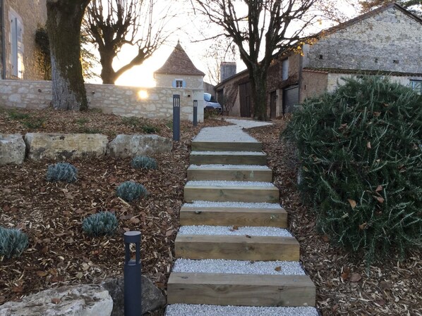 Un chemin de pierres menant à un bâtiment doté d’un toit conique, entouré d’arbres et d’arbustes.