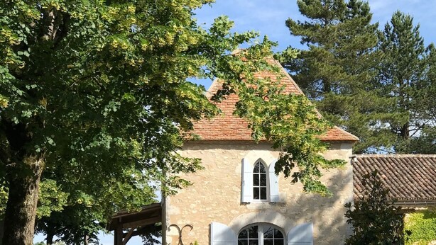 Ein Gebäude aus Stein mit einem Ziegeldach, einer überdachten Terrasse und einem großen Baum davor.