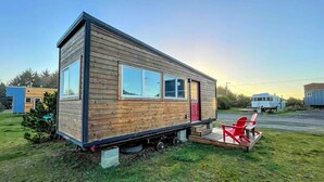 Ein modernes Mobilheim mit Flachdach, großen Fenstern und einer roten Tür. Es steht auf einer Rasenfläche, nebenan ist ein roter Stuhl zu sehen.