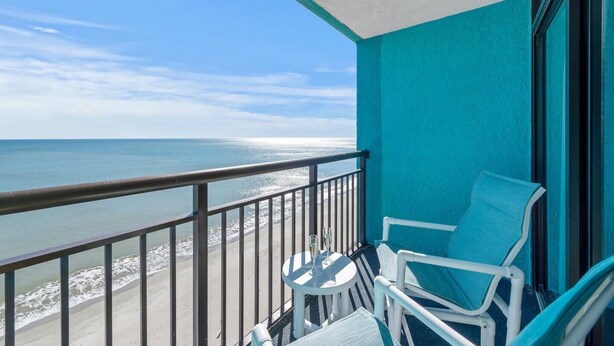 A balcony with a view of the ocean, a table with chairs, and a window with a view of the beach. - Impeccable Oceanfront Suite Top Floor (Myrtle Beach)