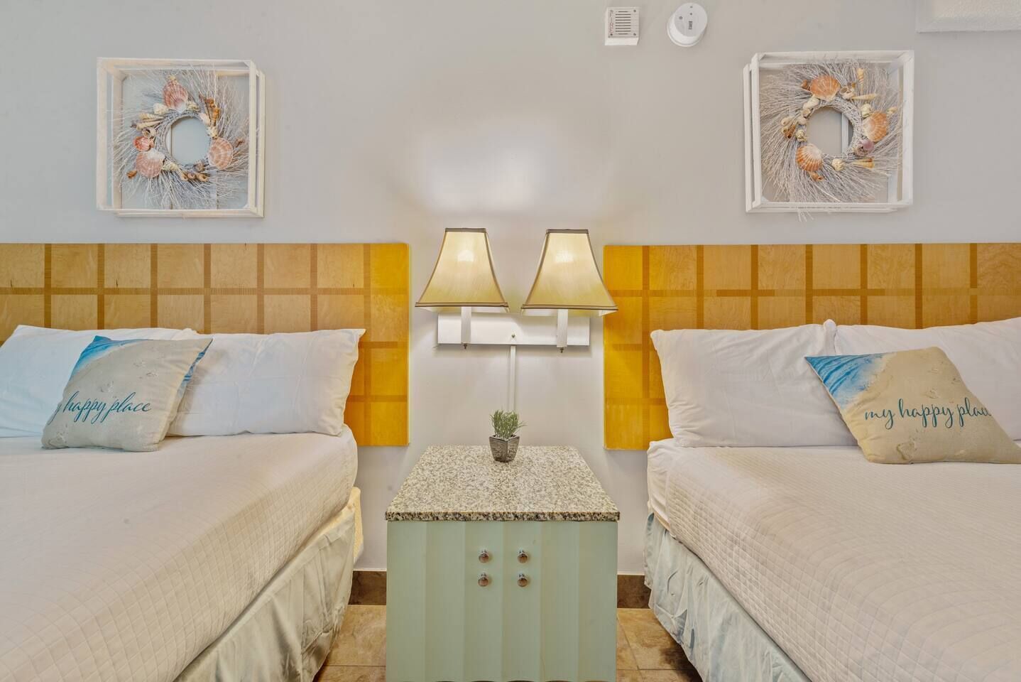 A hotel room with two beds, each with a pillow that says "happy place". There are wall-mounted lamps and two framed wall decorations.