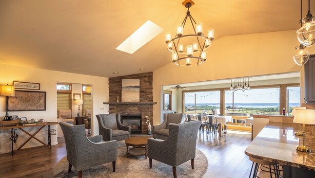 A spacious living room with a fireplace, comfortable seating, a kitchen area, and large windows offering a view.