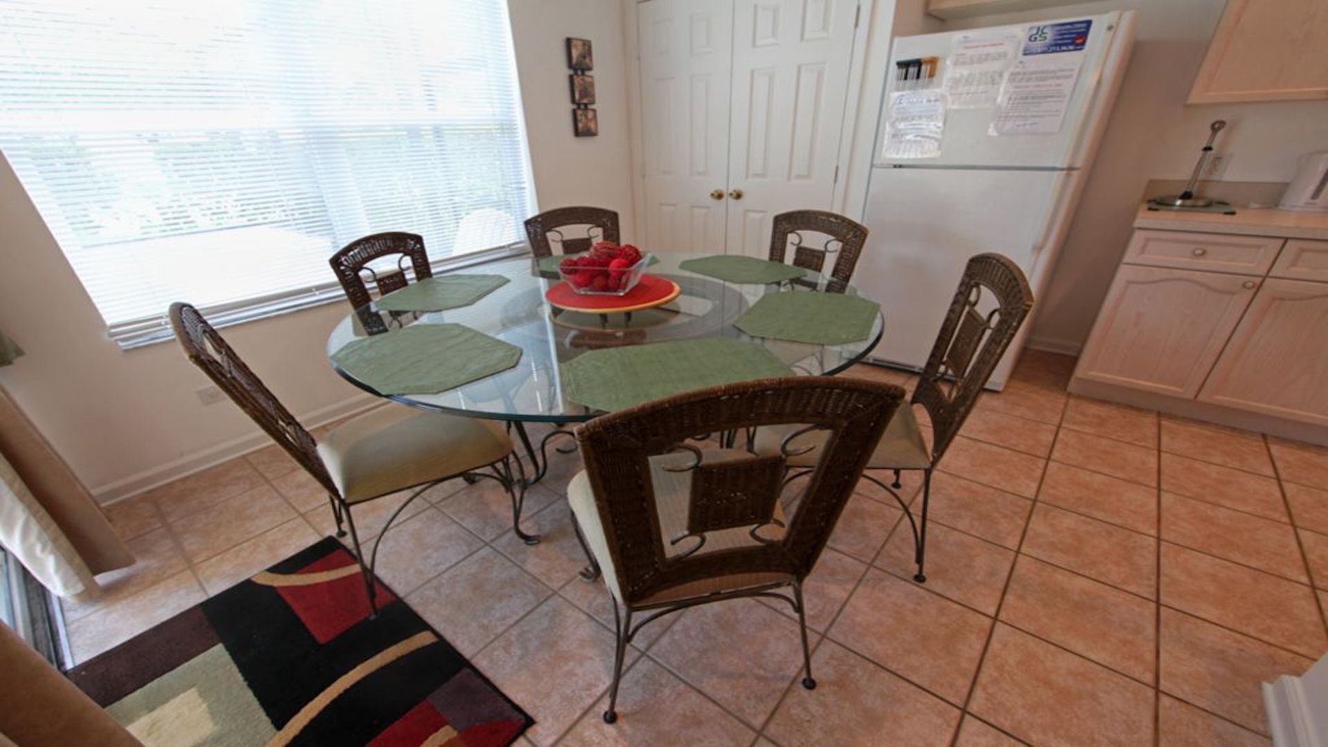 Un comedor con una mesa de vidrio rodeada de sillas de mimbre, un frutero con fruta roja, y una zona de cocina con refrigerador y armarios.