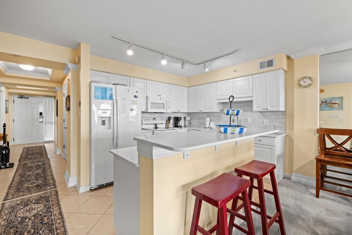 A kitchen with white cabinets, a central island with stools, a microwave, and a refrigerator.