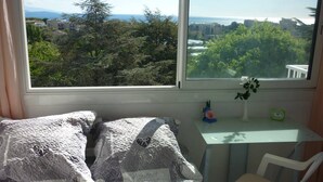 A bedroom with a large window, a bed with patterned pillows, a small table with a clock, and a chair. -  Antibes nice 3 rooms, clear and bright, beautiful sea view and swimming pool (Antibes)
