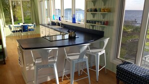 Une cuisine dotée d’un grand îlot central, de tabourets de bar blancs, d’une vue sur la mer et d’un coin repas avec des chaises bleues.