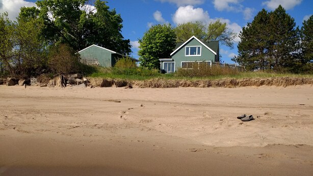 Une plage de sable où l’on voit des empreintes de pas menant à une maison verte avec une fenêtre blanche.