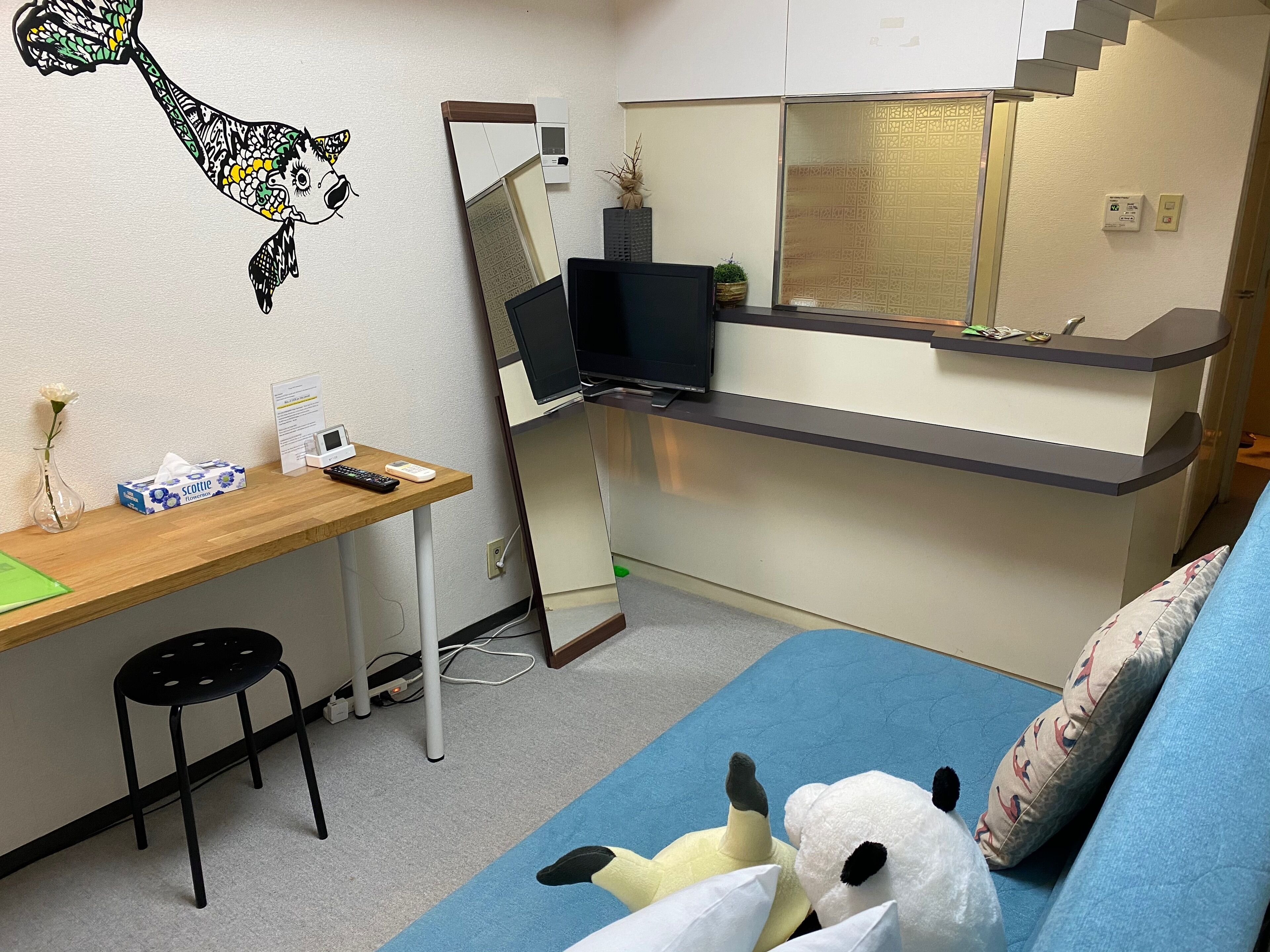A compact room with a desk, a computer, a mirror, a bed, and a small stool.