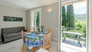 Un coin repas avec une table dressée pour quatre personnes, un canapé et un balcon offrant une vue sur les arbres et les collines.