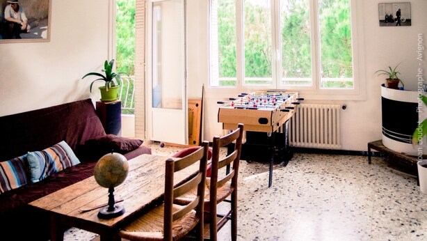 Een gezellige woonkamer met een houten tafel, een bank en een tafelvoetbaltafel. Er zijn grote ramen met uitzicht op groen buiten.