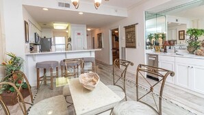 A modern kitchen with white cabinets, a central island, and a dining area with metal chairs and a marble table. - Peral 605 Sugar White Sand and Emerald Waters! Beautiful Gulf Views!  (Navarre)