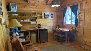 A cozy cabin interior with wooden walls, a dining table, chairs, a small refrigerator, shelves with dishes, and a television. - Ponderosa Cabin Columbia Mountain Ranch (Columbia Falls)