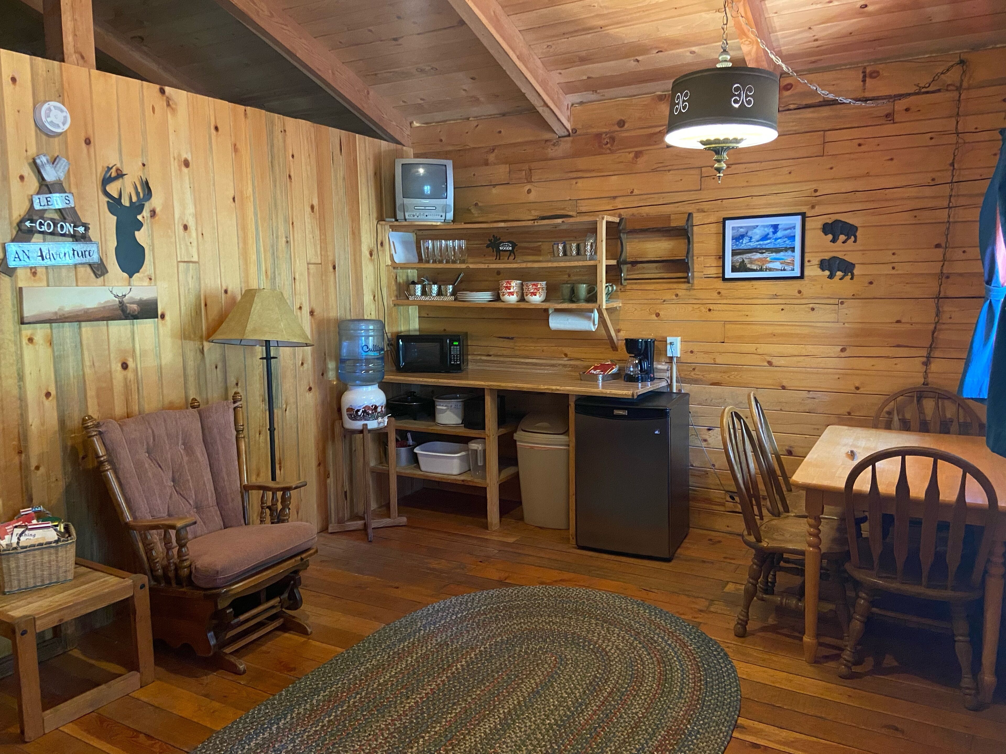 Interior acogedor de una cabaña con paredes de madera, comedor, cocina con refrigerador, microondas y cafetera, y zona de estar con una mecedora y una mesa.