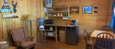 Interior acogedor de una cabaña con paredes de madera, comedor, cocina con refrigerador, microondas y cafetera, y zona de estar con una mecedora y una mesa.