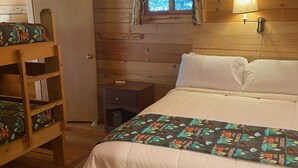 A wooden cabin bedroom with a bunk bed, a single bed, a nightstand, a lamp, and a window with curtains. - Ponderosa Cabin Columbia Mountain Ranch (Columbia Falls)