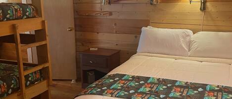 Quarto de cabana de madeira com beliche, uma cama de solteiro, mesa de cabeceira, abajur e janela com cortinas.