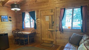 A cozy cabin interior with a wooden dining table, chairs, a sofa, and a small television. - Ponderosa Cabin Columbia Mountain Ranch (Columbia Falls)