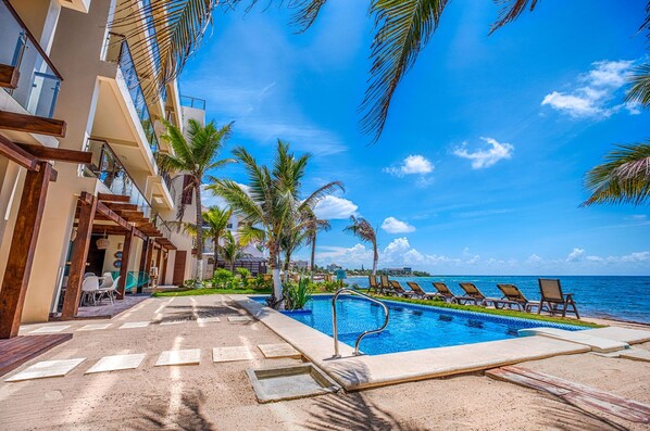 A resort pool area with a clear view of the ocean, palm trees, and a modern building with balconies.