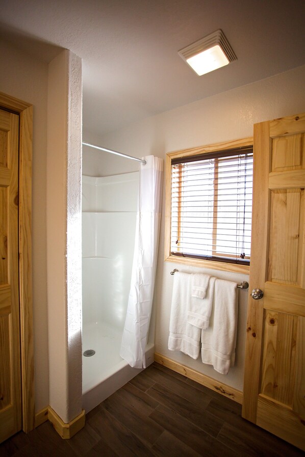 A bathroom with a glass shower enclosure, wooden doors, a window with blinds, and a towel rack.