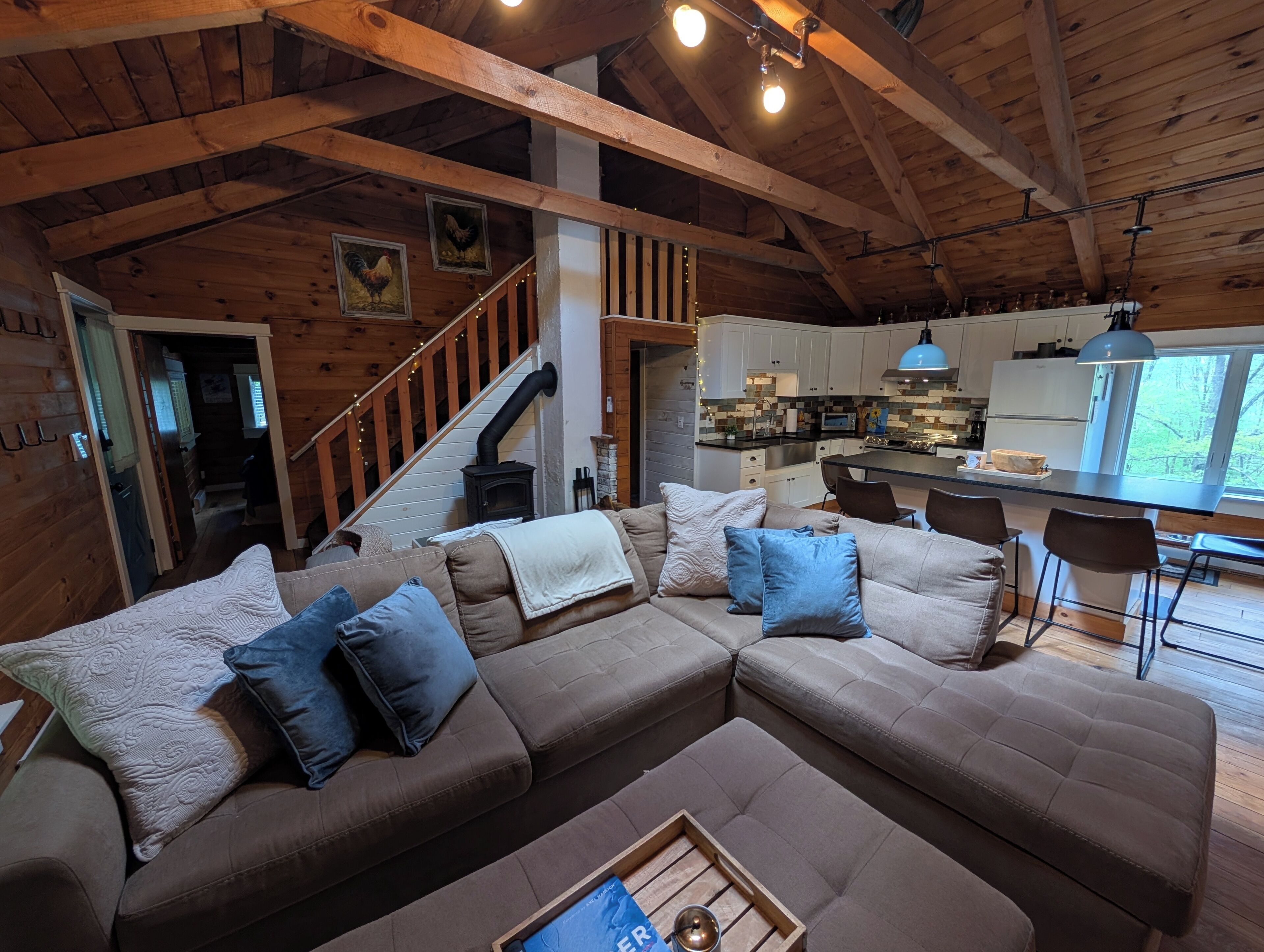 A cozy cabin interior with a large sectional sofa, a kitchen with modern appliances, and a wooden staircase.