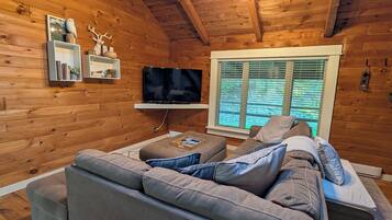 A cozy attic bedroom with wooden walls, a large gray sofa, a flat-screen TV, and a window with blinds.