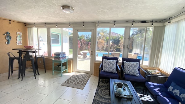 A sunlit patio with a dining table, chairs, a sofa with patterned cushions, and a view of a swimming pool and greenery outside. - Sunshine Retreat with heated Saltwater pool. Minutes to gulf beaches and IMG (Bradenton)