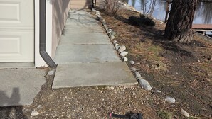 A concrete pathway leading to a garage door, flanked by a tree with pine cones and a lake in the background.