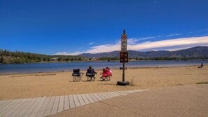 A lakeside beach with a signpost, a paved walkway, and three people sitting on beach chairs. - Cozy Creekside Retreat • Walk to Main Street • Mountain Views & Fireplace (Frisco)