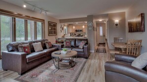 A spacious living room with a large brown leather sofa, a coffee table, and a patterned rug. There is a kitchen area in the background with modern appliances and a dining table. - Cozy Creekside Retreat • Walk to Main Street • Mountain Views & Fireplace (Frisco)