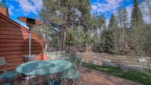 A wooden deck with a glass table and chairs, overlooking a river and forest. - Cozy Creekside Retreat • Walk to Main Street • Mountain Views & Fireplace (Frisco)