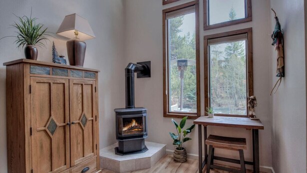A cozy room with a wood-burning stove, a cabinet with decorative glass panels, a desk with a lamp, and large windows offering a view of trees. - Cozy Creekside Retreat • Walk to Main Street • Mountain Views & Fireplace (Frisco)