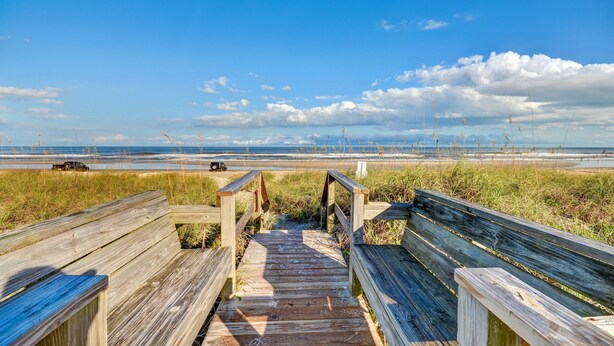 Ein hölzerner Steg führt zu einem Strand mit blauem Himmel und entfernten Wolken.
