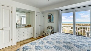 Ein Schlafzimmer mit einem großen Bett, einem Deckenventilator, einem Schminktisch, einem Stuhl und Blick auf den Strand durch eine Schiebetür.