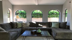 Ein Wohnzimmer mit einem Ecksofa, einem Couchtisch und drei Bogenfenstern mit Blick auf Grünflächen.