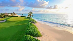 A golf course with a beach view, palm trees, and thatched-roof buildings.