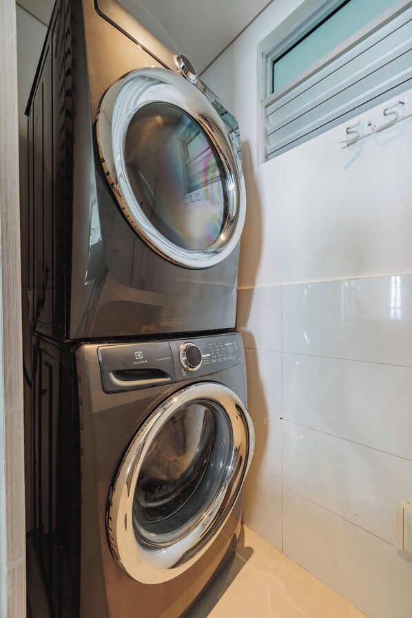 Een gestapelde wasmachine en droger in een badkamer, met een betegelde muur en een raam boven de droger.