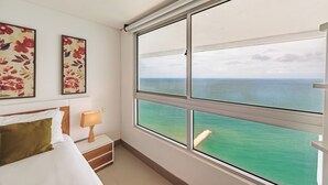 A hotel room with a large window offering a view of the sea. There are two framed floral artworks on the wall, a bed with white bedding, and a bedside table with a lamp. - Spacious & Luxury apt in Bocagrande in Palmetto Beach (Cartagena)