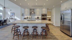 Una cocina moderna con una isla central, taburetes de bar, electrodomésticos de acero inoxidable e iluminación empotrada en el techo.