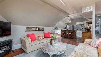 A spacious living area with a high ceiling, a sofa set, a coffee table, and a kitchen in the background.