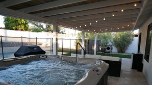 Un bain à remous doté d’un panneau de commande, entouré d’une terrasse couverte agrémentée de guirlandes lumineuses, d’une piscine et de mobilier d’extérieur.