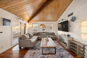 A spacious living room with a wooden ceiling, a large sectional sofa, a coffee table, and a TV on a wooden stand.